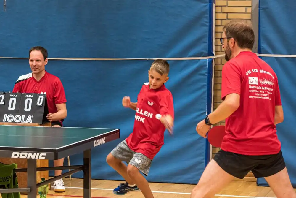 Ein Junge in einem roten Tischtennistrikot schlägt einen Tischtennisball, während ein Mann mit Brille und einem weiteren Spieler im Hintergrund auf einem Tischtennisfeld zu sehen sind.