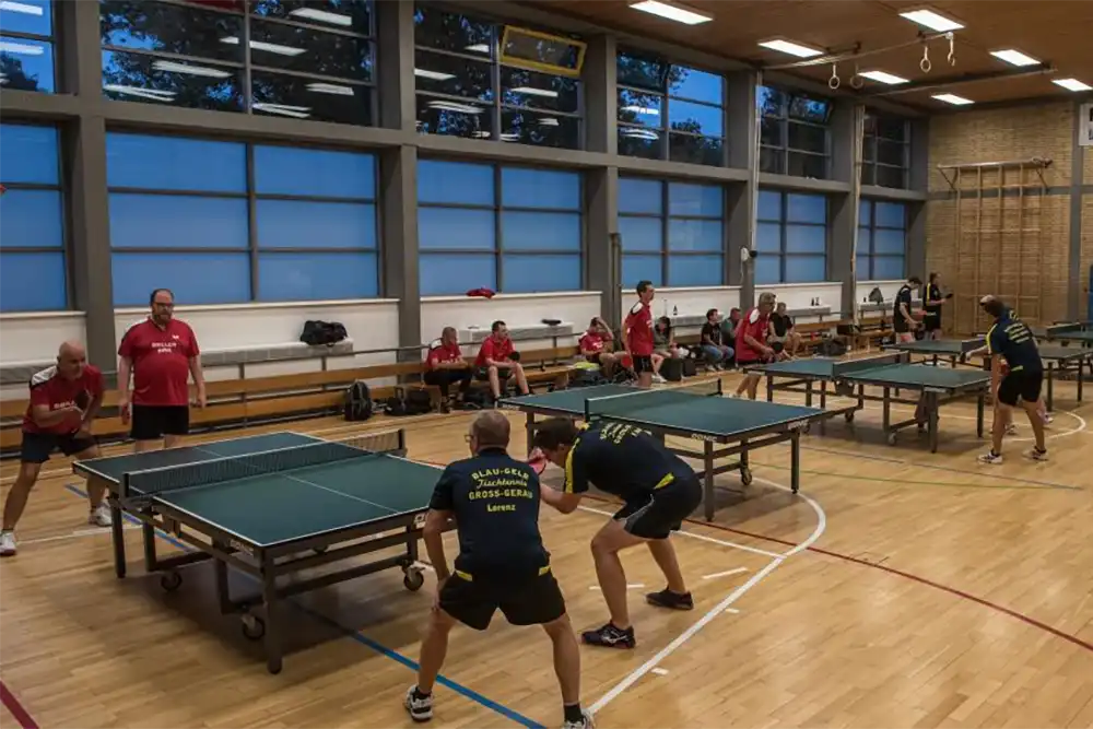 Tischtennisspieler in einer Sporthalle, zwei Spieler in schwarzer Sportkleidung und gelben Akzenten stehen an einem Tisch, bereit, den Ball zu spielen, während Zuschauer auf Bänken im Hintergrund sitzen.