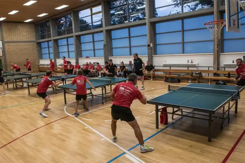 Zwei Tischtennisspieler in roten Trikots spielen in einer Sporthalle, während Zuschauer auf Bänken sitzen.