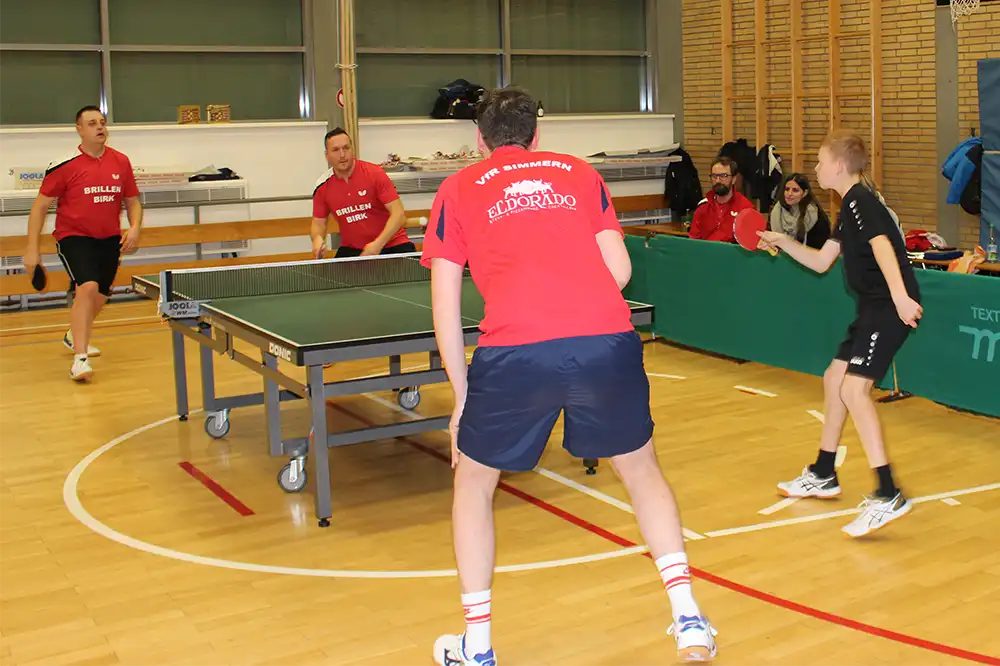 Zwei Tischtennisspieler in roten Trikots stehen an einem Tischtennis-Tisch, während zwei andere Spieler sich auf den nächsten Schlag vorbereiten. Im Hintergrund sind Zuschauer zu sehen.