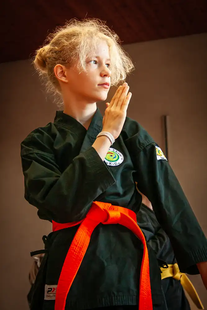 Ein junges Mädchen in einem schwarzen Hapkido-Anzug mit einem orangefarbenen Gürtel, das eine Kampfkunstpose einnimmt. Sie hat blonde Haare und schaut konzentriert nach vorne, während sie eine Hand in einer verteidigenden Geste hebt.