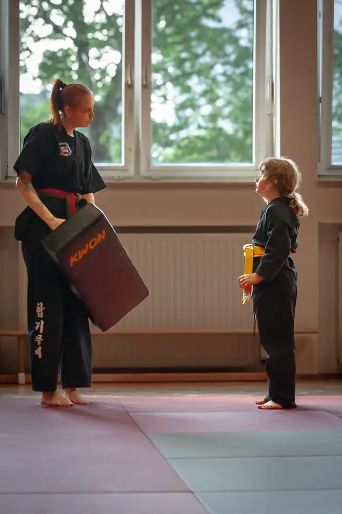 Übungsleiterin Maxime Sonnet in schwarzem Hapkido-Anzug mit rotem Gürtel hält ein Trainingspolster in der Hand, während ein Mädchen mit einem gelben Gürtel ihr gegenübersteht und aufmerksam zuhört.