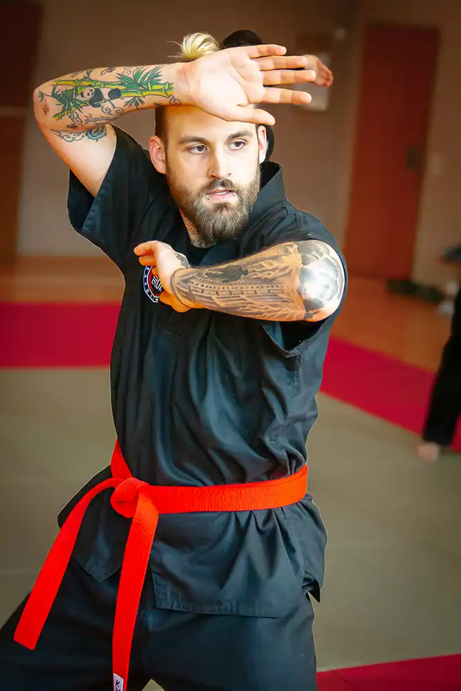 Ein Mann in schwarzer Kampfkunstkleidung mit einem orangefarbenen Gürtel zeigt eine Hapkido-Technik. Seine Arme sind in einer Abwehrhaltung positioniert, während er konzentriert in die Kamera blickt. Tätowierungen sind auf seinen Armen sichtbar. Im Hintergrund sind unscharfe Silhouetten anderer Personen in einer Kampfkunstschule zu erkennen.