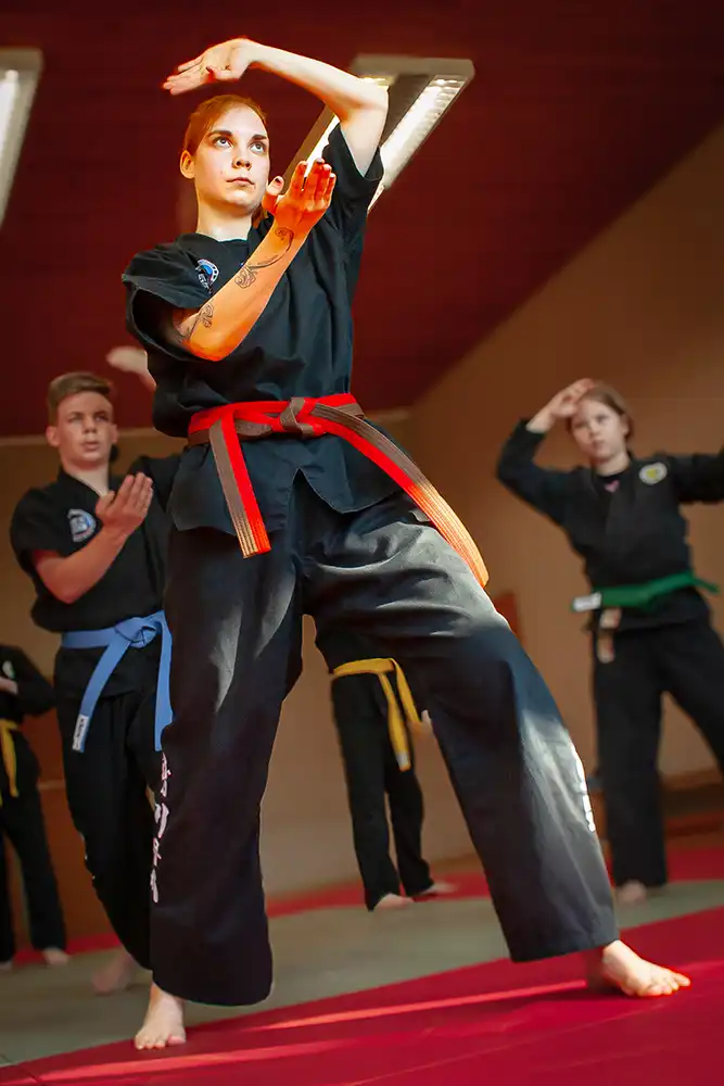 Eine Kampfsportübung in einer Hapkido-Trainingseinheit, in der eine Frau mit rotem Gürtel eine Technik demonstriert, während andere Teilnehmer mit blauen und gelben Gürteln im Hintergrund üben.