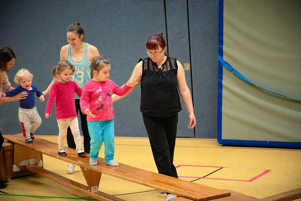 Eine Gruppe von Kindern, die mit Erwachsenen auf einem Barren balanciert. Die Kinder tragen bunte Oberteile und Hosen. Die Erwachsenen unterstützen die Kinder beim Gehen auf der Bank. Eine Szene aus dem Eltern-Kind-Turnen des VfR Simmern.