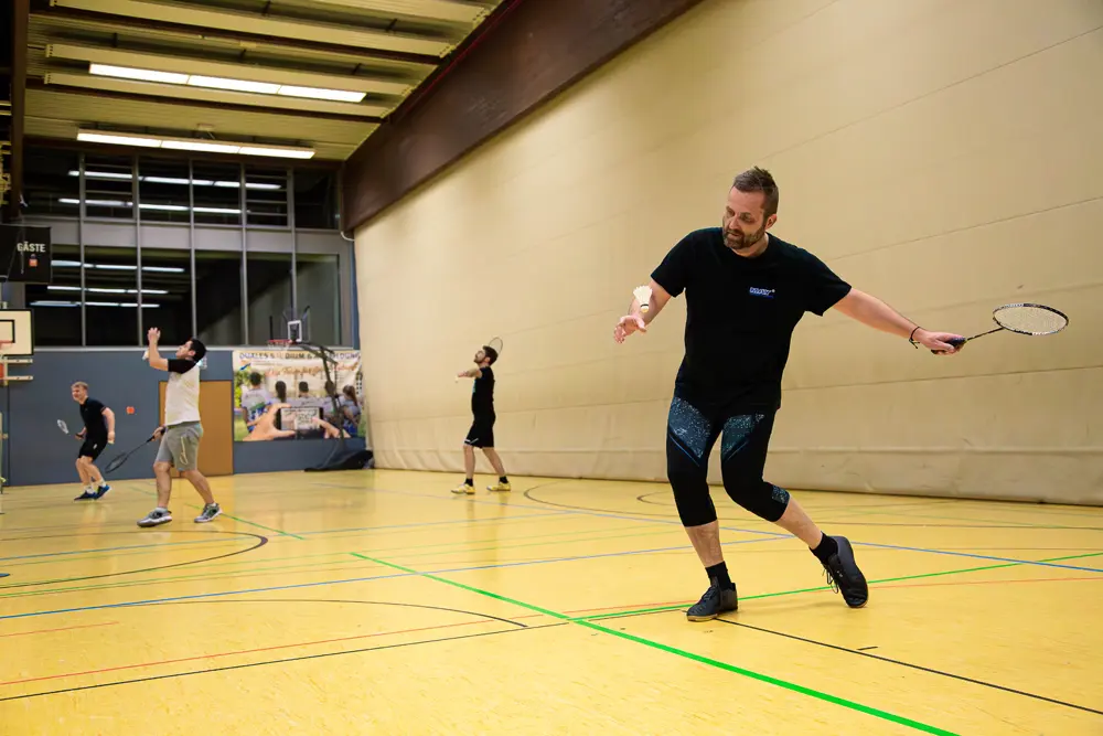 Eine Gruppe von vier Männern spielt Badminton in einer Sporthalle. Ein Spieler in schwarzem T-Shirt und Leggings führt einen Schlag mit dem Badmintonschläger aus, während die anderen Spieler sich im Hintergrund bewegen. Die Halle hat einen Holzfußboden und eine helle Wand.