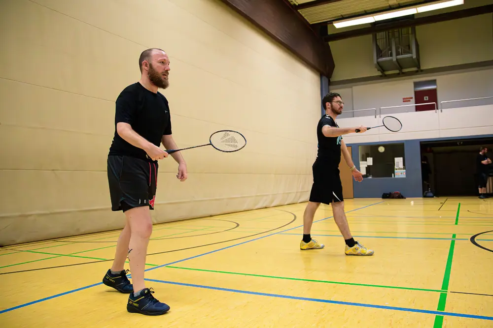Zwei Männer spielen Badminton in einer Sporthalle. Der Mann auf der linken Seite hat einen Bart und hält einen Badmintonschläger in der Hand, während der andere Mann auf der rechten Seite ebenfalls einen Schläger hält und sich in einer spielerischen Pose befindet. Der Boden ist gelb mit grünen Linien für das Badmintonfeld.