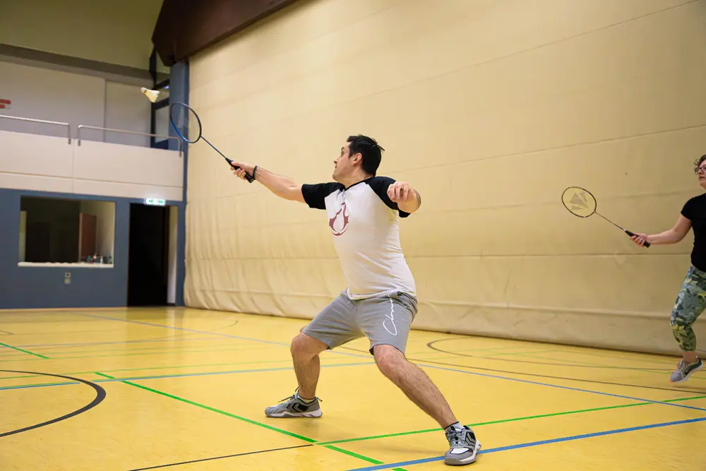 Ein Mann spielt Badminton in einer Sporthalle, während er einen Schläger in der Hand hält und einen Federball schlägt. Im Hintergrund ist eine weitere Spielerin sichtbar, die ebenfalls aktiv am Spiel teilnimmt.