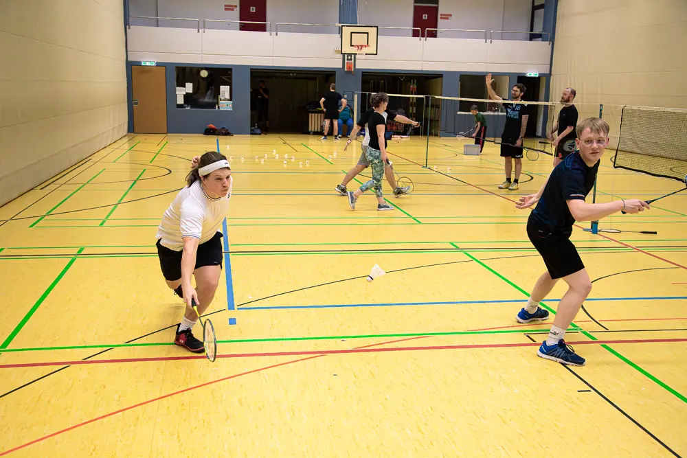 Eine Badminton-Trainingseinheit in einer Sporthalle, bei der mehrere Spieler aktiv sind. Eine Spielerin in einem weißen Shirt und einem Stirnband schlägt mit einem Schläger einen Federball, während ein weiterer Spieler in schwarzer Kleidung bereit ist, zu spielen. Die Halle hat ein gelb gefärbtes Spielfeld mit klaren Linien und Badmintonnetzen im Hintergrund.