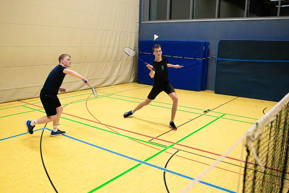 Zwei junge Männer spielen Badminton in einer Sporthalle. Der Spieler auf der linken Seite führt einen Aufschlag aus, während der Spieler auf der rechten Seite sich in einer Angriffshaltung befindet. Beide tragen Sportbekleidung und Schläger. Der Boden ist gelb mit bunten Linien für das Spielfeld.