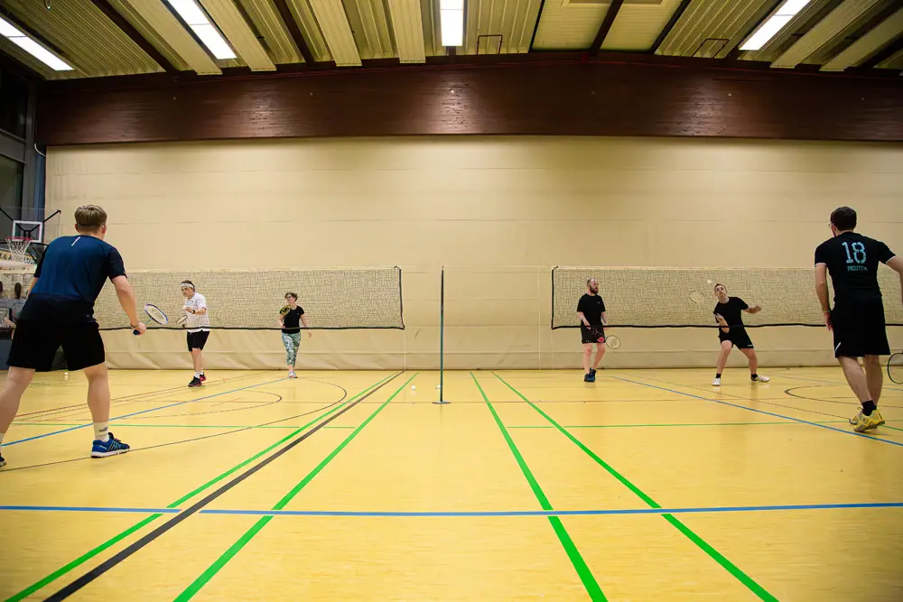 Eine Gruppe von fünf Personen spielt Badminton in einer Sporthalle. Die Spieler stehen auf einem gelben Hallenboden mit grünen Linien und sind in zwei Teams aufgeteilt, die sich gegenüberstehen. Im Hintergrund sind Badmintonnets und ein Netz sichtbar.