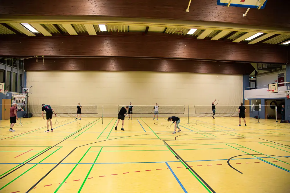 Badminton-Spieler in einer Sporthalle beim Training, mit mehreren Spielfeldern und Netzen im Hintergrund.