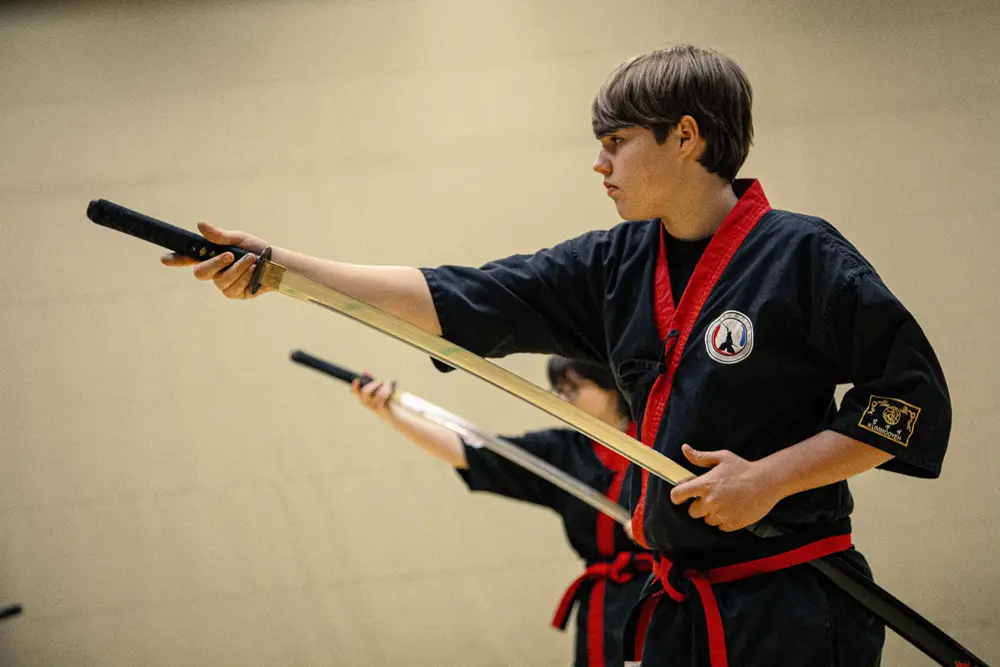 Ein junger Kämpfer in traditioneller koreanischer Kampfkunstkleidung hält ein Schwert in der Kummooyeh-Pose, während er konzentriert auf einen anderen Kämpfer im Hintergrund schaut.
