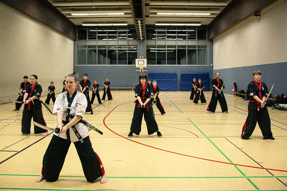 Eine Gruppe von Praktizierenden der koreanischen Kampfkunst Kummooyeh steht in einer Sporthalle. Sie tragen schwarze Trainingsanzüge mit roten Akzenten und halten traditionelle Schwerter. Die Szene zeigt eine Mischung aus Männern und Frauen, die konzentriert und in einer synchronisierten Haltung für das Training bereit sind.