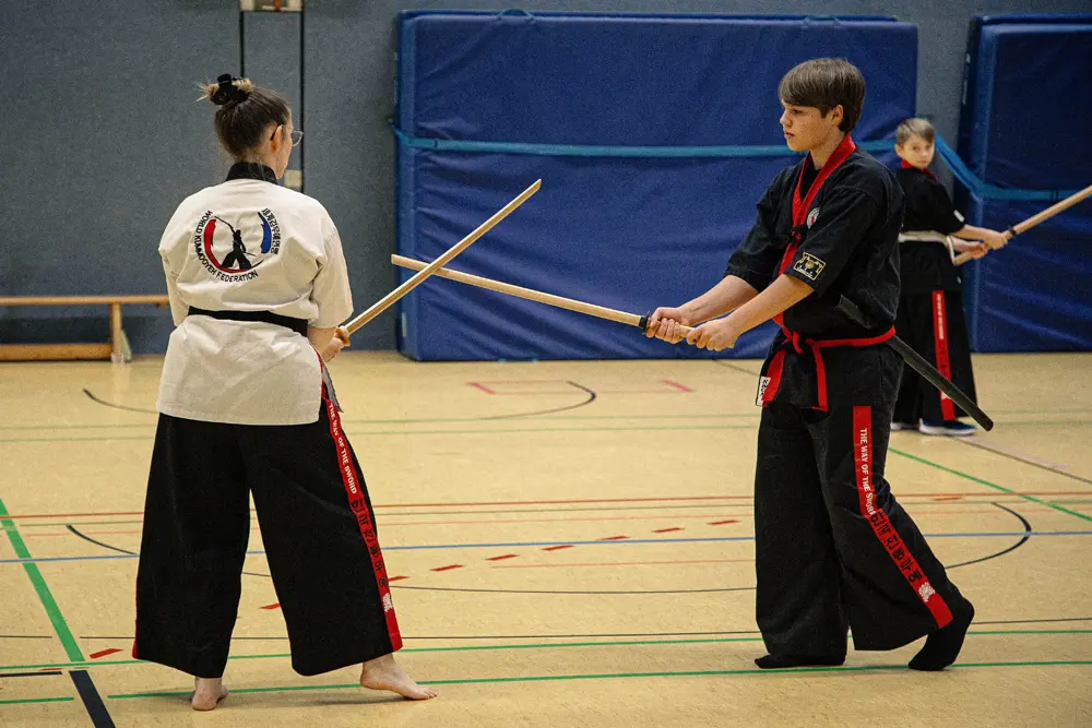 Abteilungsleiterin Silke Theis übt mit einem Kummooyeh, eine koreanische Kampfkunst, in einer Sporthalle. Der Junge trägt ein schwarzes Karate-Outfit mit rotem Gürtel, während Silke Theis ein weißes Oberteil mit schwarzem Gürtel trägt. Beide halten Holzschwerter und stehen in einer Kampfhaltung.