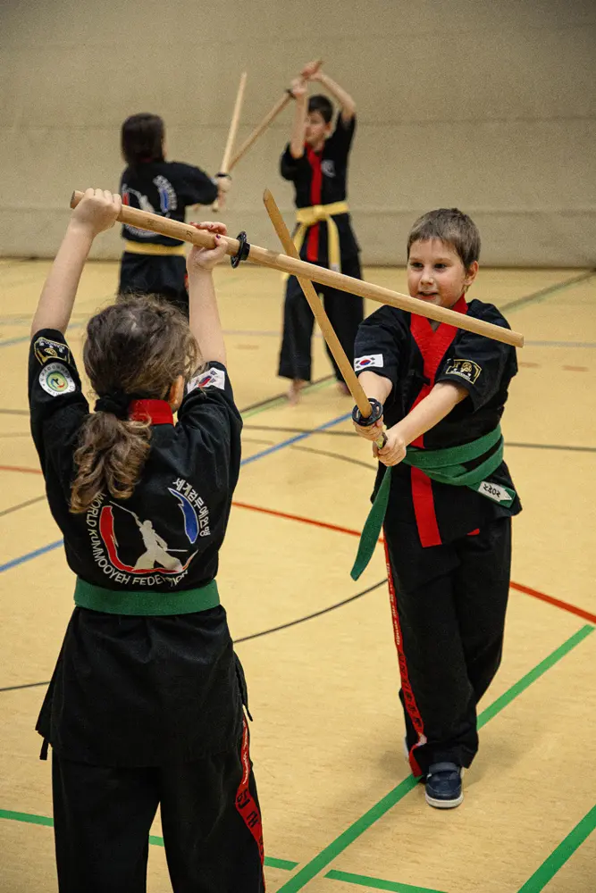 Kinder in schwarzer Kampfkunstbekleidung üben Kummooyeh mit Holzstöcken in einer Sporthalle. Einige tragen grüne und gelbe Gürtel.