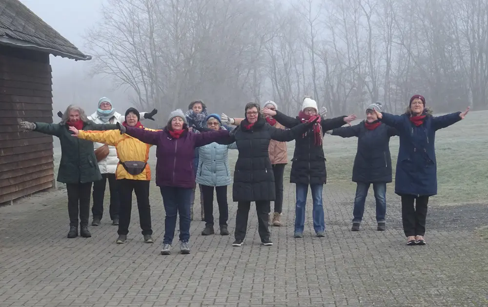Eine Gruppe von Frauen steht im Freien in einer Reihe und führt Armbewegungen aus, während sie in warmer Kleidung und Schals gekleidet sind. Die Szene findet in einem nebligen Umfeld statt, was eine ruhige Atmosphäre schafft.