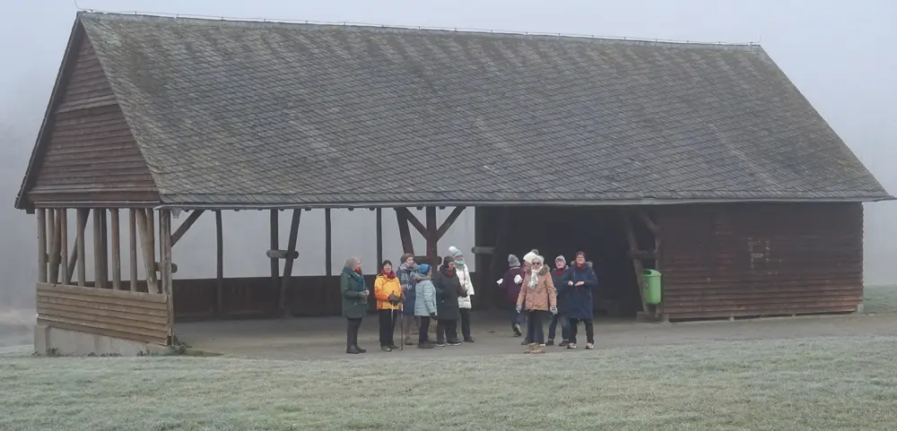 Eine Gruppe von Frauen steht unter einem überdachten Holzbau in einem nebligen Umfeld, während sie sich auf einen Walking-Treff vorbereiten. Sie tragen verschiedene Farben und Bekleidungen, die für die kalte Witterung geeignet sind.