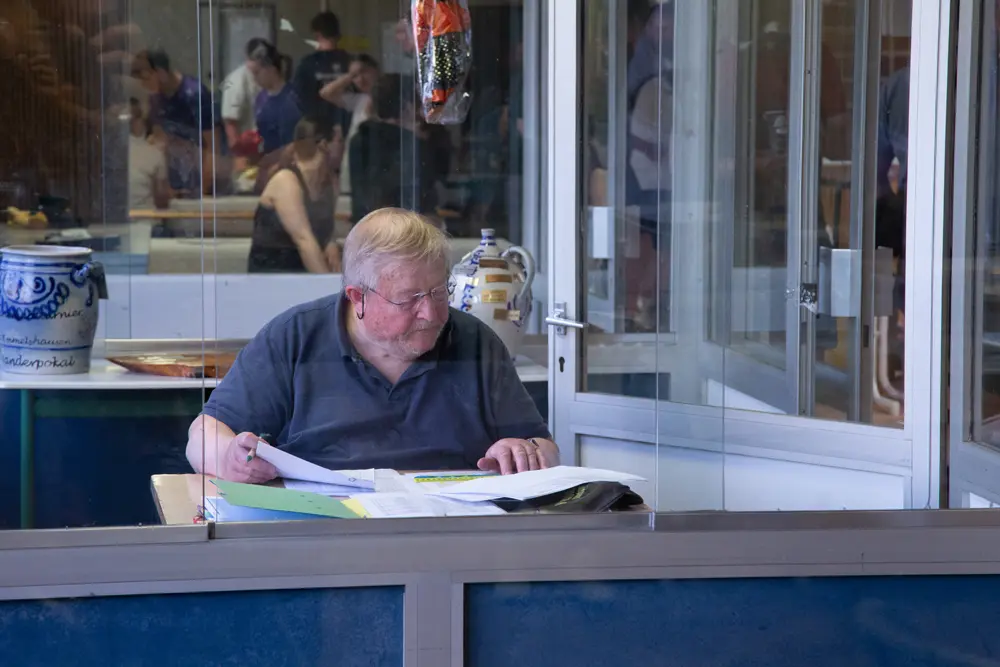 Zwei junge Männer spielen Badminton in einer Sporthalle. Der Spieler auf der linken Seite führt einen Aufschlag aus, während der Spieler auf der rechten Seite sich in einer Angriffshaltung befindet. Beide tragen Sportbekleidung und Schläger. Der Boden ist gelb mit bunten Linien für das Spielfeld.