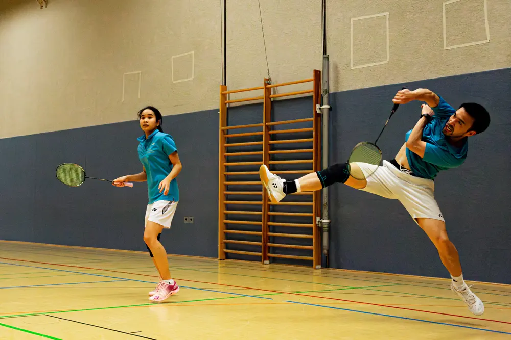 Eine Gruppe von fünf Personen spielt Badminton in einer Sporthalle. Die Spieler stehen auf einem gelben Hallenboden mit grünen Linien und sind in zwei Teams aufgeteilt, die sich gegenüberstehen. Im Hintergrund sind Badmintonnets und ein Netz sichtbar.