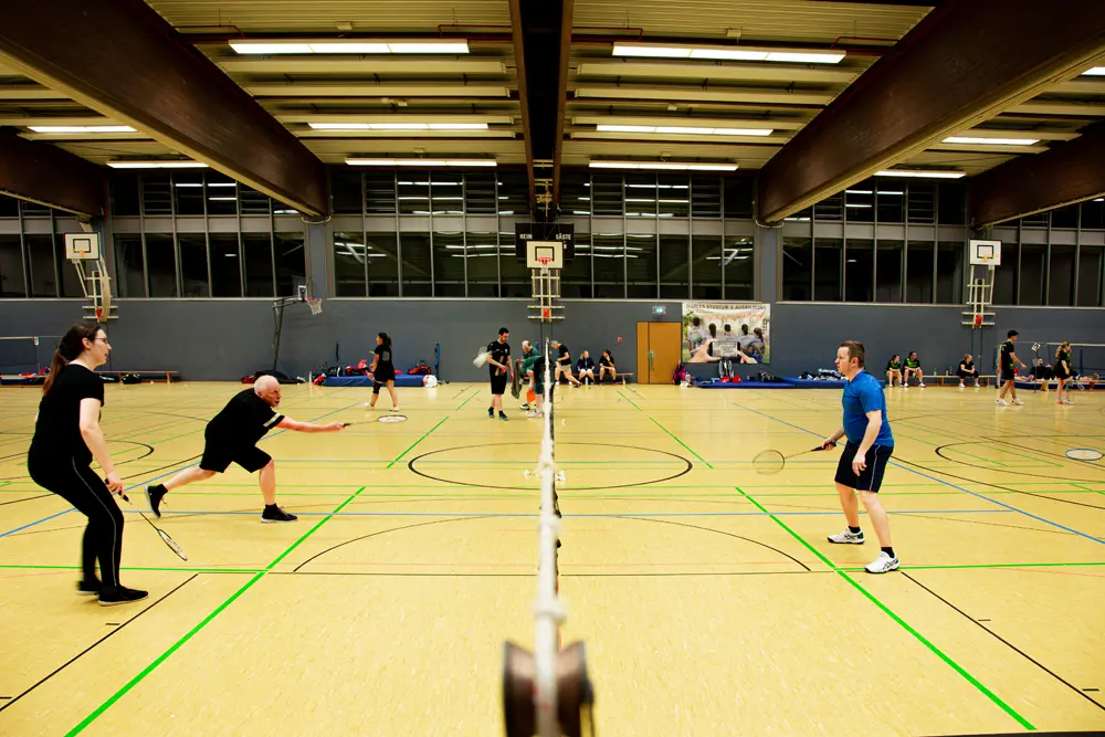 Zwei Männer spielen Badminton in einer Sporthalle. Der Mann auf der linken Seite hat einen Bart und hält einen Badmintonschläger in der Hand, während der andere Mann auf der rechten Seite ebenfalls einen Schläger hält und sich in einer spielerischen Pose befindet. Der Boden ist gelb mit grünen Linien für das Badmintonfeld.