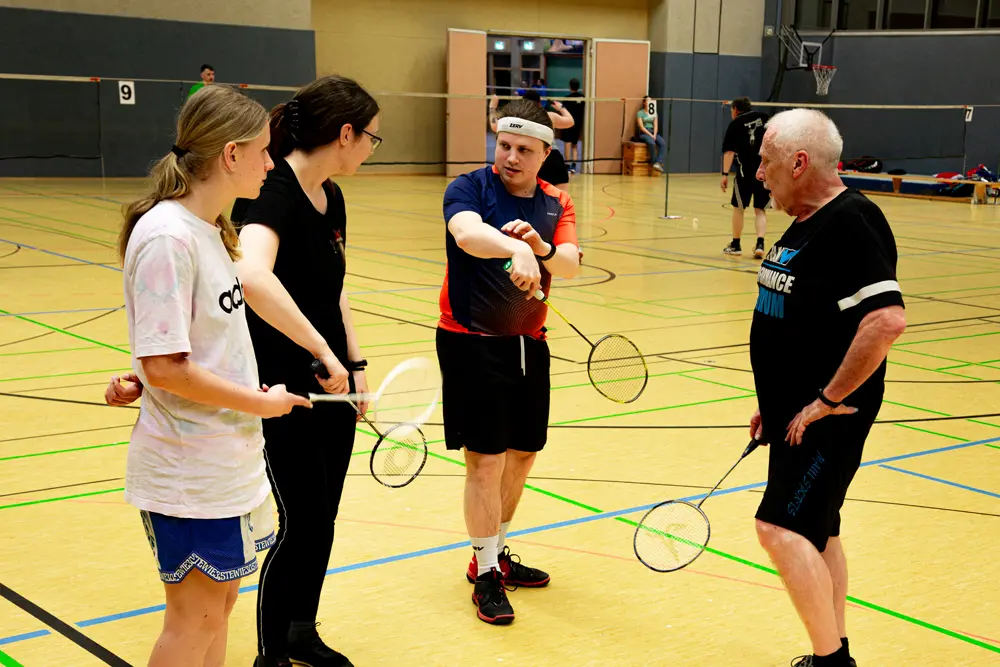 Eine Badminton-Trainingseinheit in einer Sporthalle, bei der mehrere Spieler aktiv sind. Eine Spielerin in einem weißen Shirt und einem Stirnband schlägt mit einem Schläger einen Federball, während ein weiterer Spieler in schwarzer Kleidung bereit ist, zu spielen. Die Halle hat ein gelb gefärbtes Spielfeld mit klaren Linien und Badmintonnetzen im Hintergrund.