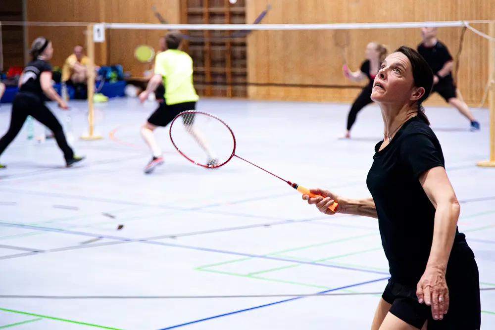 Badminton-Spieler in einer Sporthalle beim Training, mit mehreren Spielfeldern und Netzen im Hintergrund.