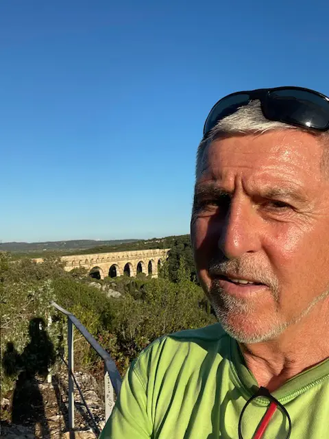 Portrait von Johannes Klein, Abteilungsleiter der Triathlonabteilung des VfR Simmern, mit grauen Haaren und kurzem Bart, steht im grünen Hemd und mit einer Sonnenbrille auf dem Kopf im Freien, hinter sich ein steinernes Aquädukt und eine üppige Landschaft unter einem klaren blauen Himmel.