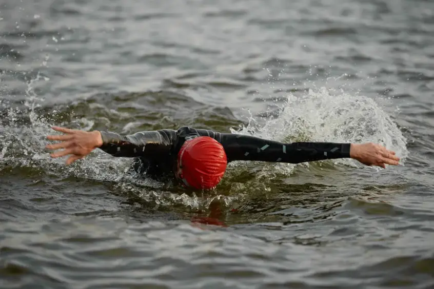 Ein Schwimmer der Triathlonabteilung des VfR Simmern, der eine rote Badekappe und einen schwarzen Neoprenanzug trägt, führt Freistilzüge im offenen Wasser aus, wobei er die Arme ausstreckt und von Spritzern umgeben ist.