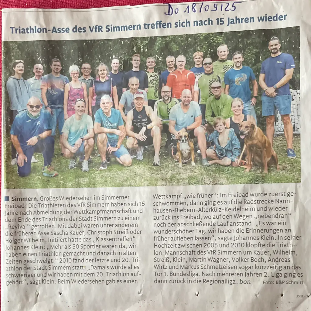 Eine große Gruppe von Männern und Frauen in Sportkleidung lächelt im Freien für ein Triathlon-Meeting-Foto, stehend und kniend auf einer Wiese mit Bäumen hinter ihnen. Hier der Zeitungsartikel aus der Rhein Hunsrück Zeitung vom 18. September 2025 über ihr Wiedersehen.