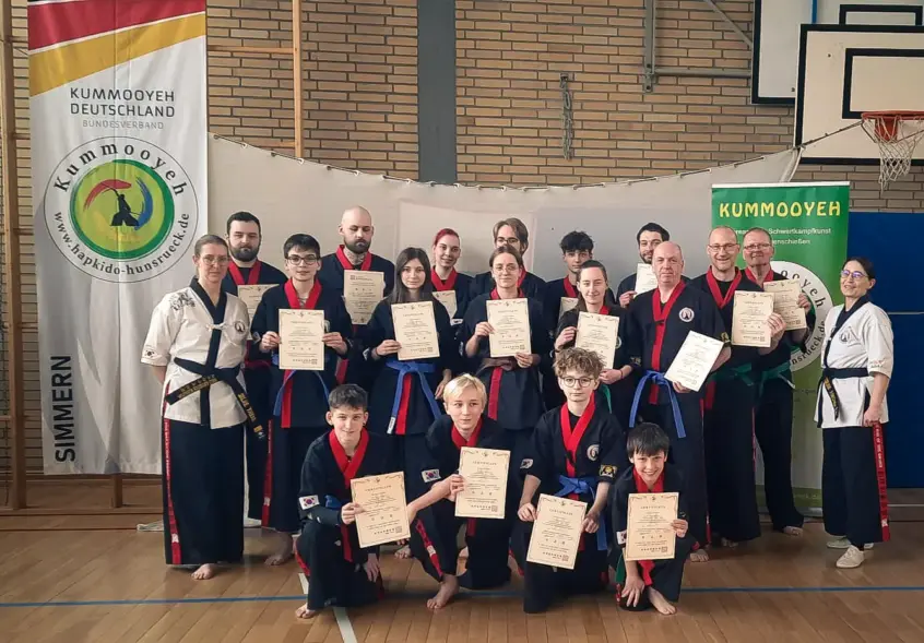 Gruppe von Kummooyeh-Prüfungsteilnehmern, die stolz ihre Zertifikate nach bestandener Gürtelprüfung halten. Die Teilnehmer sind in traditionellen schwarz-roten Kampfkünstler-Outfits gekleidet. Im Hintergrund sind Banner des Kummooyeh Deutschland Bundesverbands zu sehen, und die Umgebung ist eine Sporthalle mit Holzfußboden.