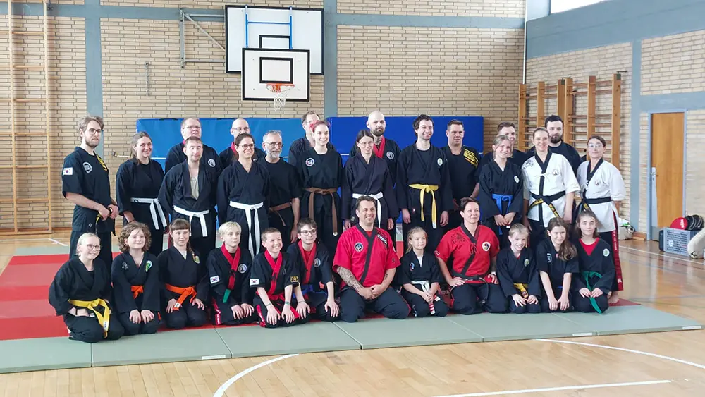 Eine Gruppe von Kampfsportlern, darunter Großmeister Frankie Lacroix und Cindy Lacroix, steht in einem Sportraum in der VfR Simmern. Die Teilnehmer tragen verschiedene Gürtel, darunter schwarz, rot und gelb, und sind in traditionellen Trainingsanzügen gekleidet. Im Hintergrund sind Sportgeräte und eine Basketball-Anlage sichtbar. Die Gruppe posiert gemeinsam zum Foto.