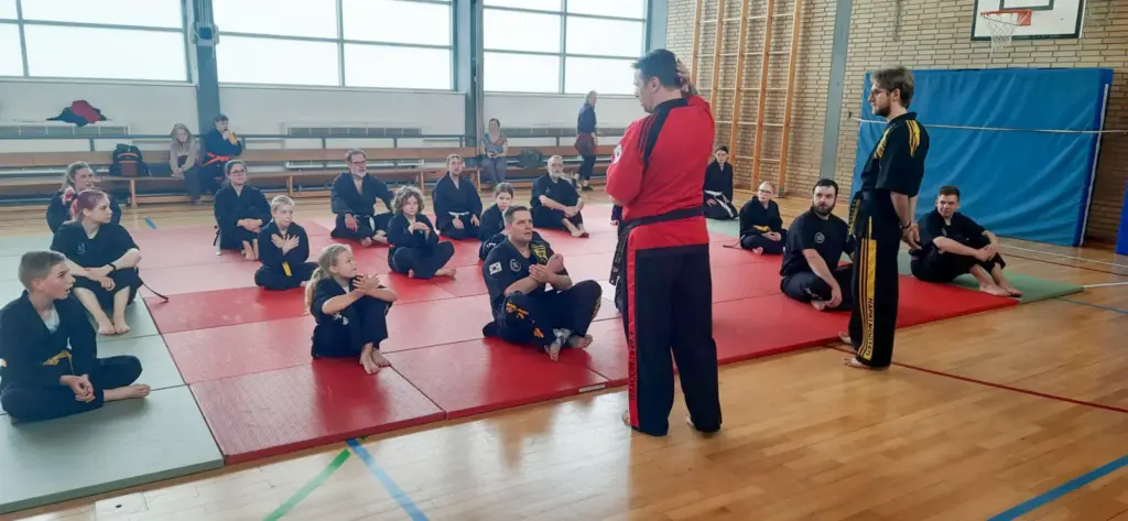 Eine Trainingssitzung im Kummooyeh unter der Leitung von Großmeister Frankie Lacroix und Cindy Lacroix. Die Teilnehmer sitzen auf Matten in einer Sporthalle, während einer der Trainer (in rotem Anzug) spricht. Im Hintergrund sind weitere Teilnehmer zu sehen, die in schwarzen Trainingsanzügen gekleidet sind. Die Atmosphäre ist konzentriert und lehrreich, ideal für das Training im VfR Simmern.