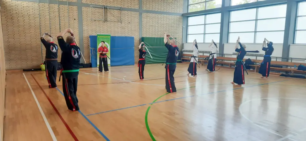 Eine Gruppe von Kummooyeh-Trainierenden des VfR Simmern in der Turnhalle der Rottmannschule. Die Teilnehmer tragen Trainingsanzüge mit dem Logo von Großmeister Frankie Lacroix und Cindy Lacroix. Sie stehen in einer Reihe, heben ihre Klingen über den Kopf und führen synchronisierte Bewegungen aus. Im Hintergrund sind Trainingsmaterialien und Bänke sichtbar.