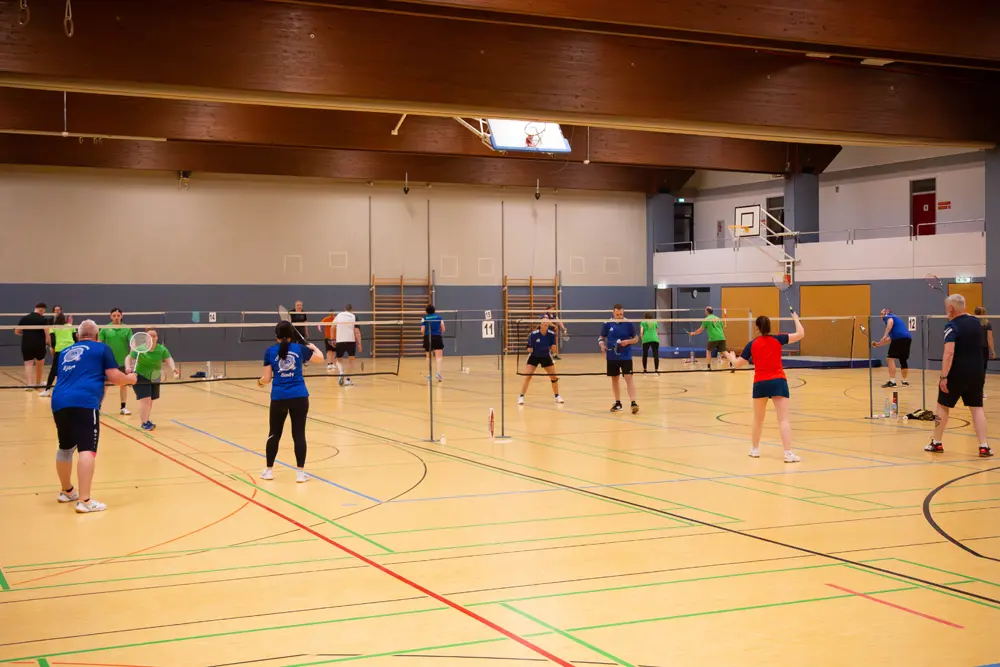 Badmintonspiel während des Hexennachtturniers 2026 in der Halle des VfR Simmern. Mehrere Spieler in blauen und grünen Trikots sind aktiv am Spielen, während sich das Spielfeld mit Netzen und Linien zeigt. Es sind sowohl Einzel- als auch Mixed-Matches zu sehen, in einer freundlichen Wettkampfatmosphäre.
