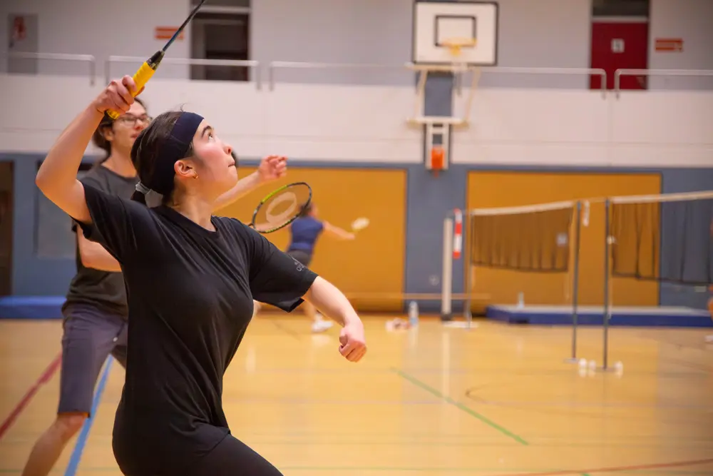 Eine Badmintonspielerin in einem Sportraum hebt ihren Schläger mit einer dynamischen Bewegung, während sie sich auf den Ball konzentriert. Im Hintergrund sind weitere Spieler, die ebenfalls Badminton spielen. Die Atmosphäre ist lebhaft und sportlich, passend zum Hexennachtturnier 2026 des VfR Simmern im Mixed-Format.