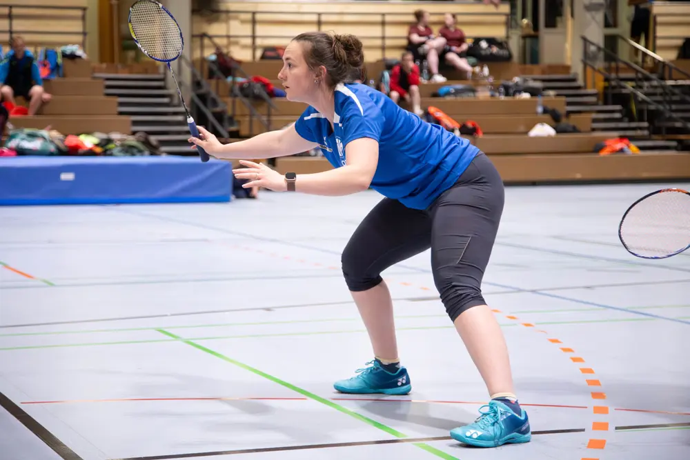 Eine Badmintonspielerin in einem blauen Trikot und dunklen Leggings steht in einer dynamischen Position auf dem Spielfeld. Sie hält einen Badmintonschläger in der rechten Hand und bereitet sich darauf vor, den Ball zu schlagen. Im Hintergrund sind Zuschauer auf Tribünen zu sehen, während die Halle für das Hexennachtturnier 2026 des VfR Simmern vorbereitet wird.