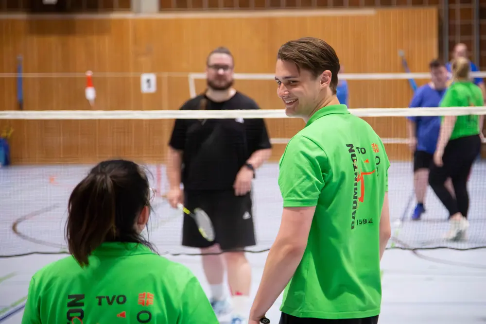 Eine Gruppe von Badmintonspielern, darunter ein Mann in einem grünen Shirt mit dem Aufdruck "Badminton" und einem Logo, lächelt und spricht mit einer Frau, die ebenfalls ein grünes Shirt trägt. Im Hintergrund sind weitere Spieler in blauen und schwarzen Shirts zu sehen, die sich auf einem Badmintonfeld in einer Sporthalle für das Hexennachtturnier 2026 des VfR Simmern vorbereiten. Die Atmosphäre ist freundlich und sportlich, mit Badmintonschlägern und einem Netz sichtbar.