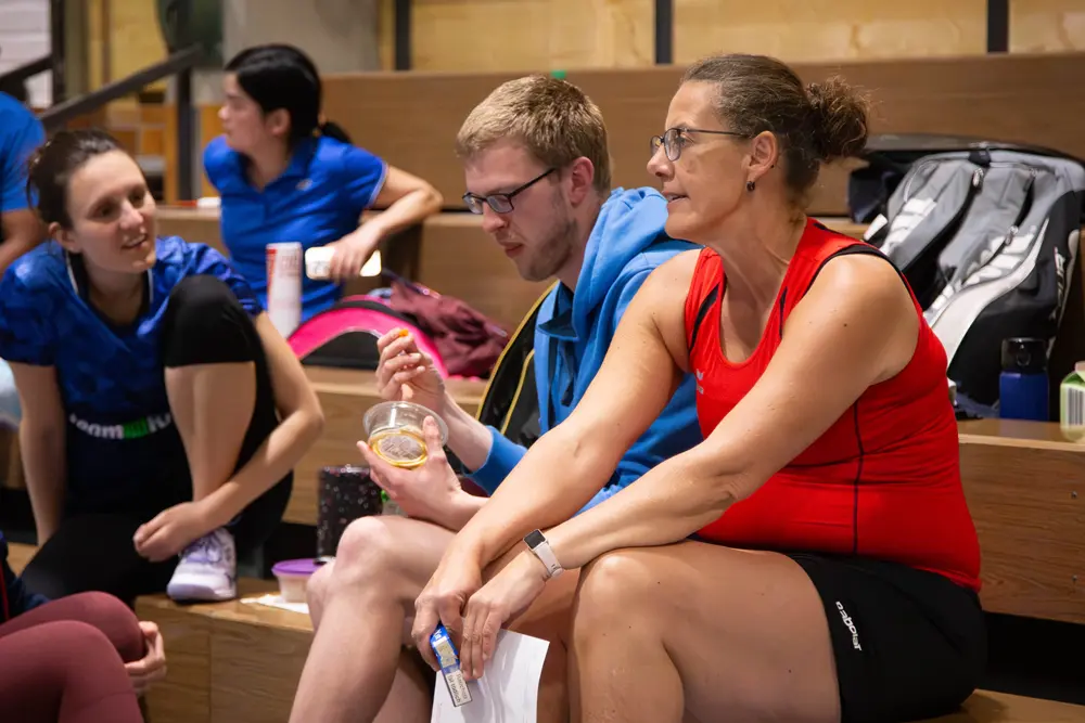 Drei Badmintonspieler sitzen auf einer Holzbank. Melanie Wies in rotem Trikot und Shorts spricht mit einem Mann in blauer Jacke, der einen Snack in einer Schüssel hält. Im Hintergrund sind weitere Spielerinnen in blauen Trikots zu sehen. Das Bild vermittelt eine entspannte Atmosphäre während des Hexennachtturniers 2026 des VfR Simmern.