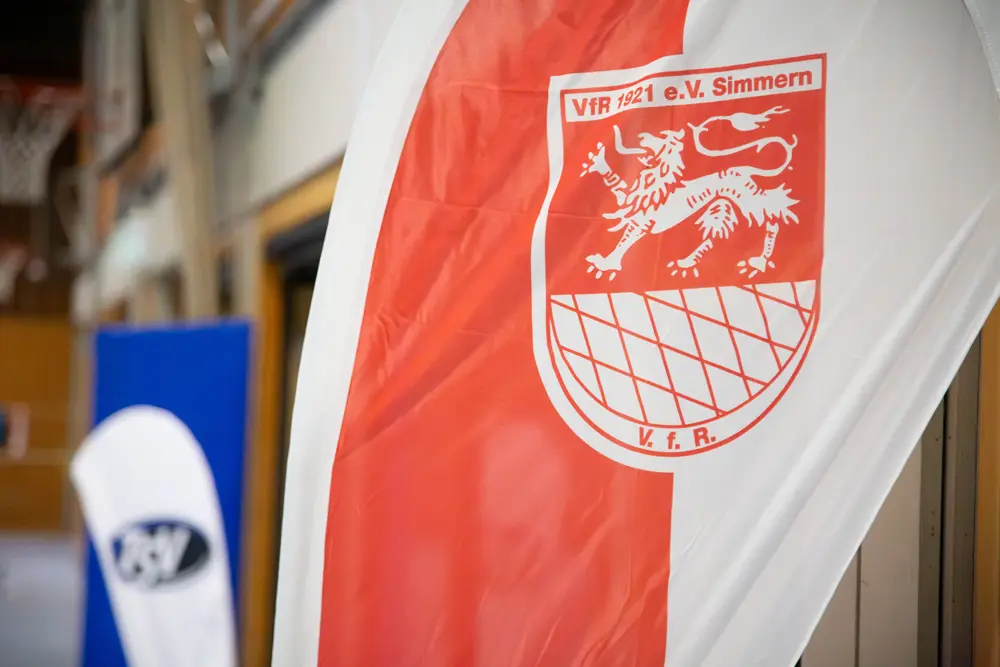 Flagge des VfR Simmern, die in einer Sporthalle hängt. Die Flagge zeigt das Wappen des Vereins mit einem stilisierten Löwen und dem Schriftzug "VfR 1921 e.V. Simmern". Im Hintergrund ist das Banner des TSV Emmelshausen zu sehen