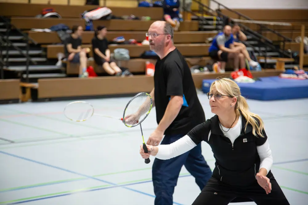 Ein Badmintonspiel während des Hexennachtturnier des VfR Simmern 2026. Zu sehen sind ein Mann und eine Frau in einer Sporthalle. Die Frau, mit langen blonden Haaren und Brille, bereitet sich darauf vor, den Federball zu schlagen, während der Mann hinter ihr steht und aufmerksam zuschaut. Im Hintergrund sind weitere Zuschauer auf den Tribünen zu sehen.