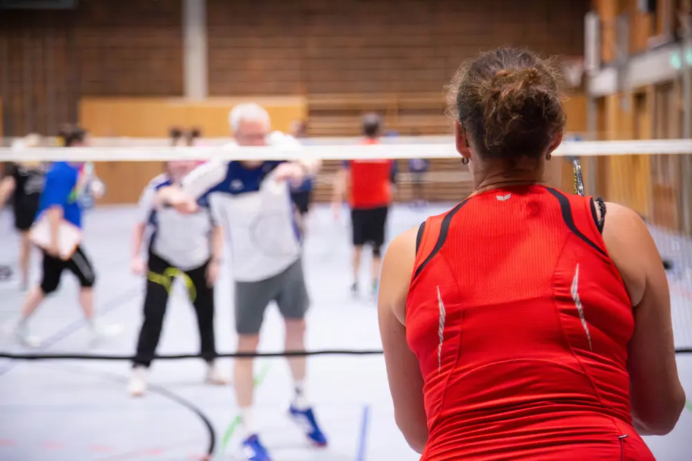Eine Gruppe von Badmintonspielern in einer Sporthalle, die an einem Mixed-Turnier teilnehmen. Im Vordergrund ist Melanie Wies zu sehen in einem roten Sporttop, die auf den Aufschlag wartet. Im Hintergrund sind mehrere Spieler in unterschiedlichen Sportoutfits, die an den Spielen des 36. Hexennachtturnier des VfR Simmern teilnehmen.