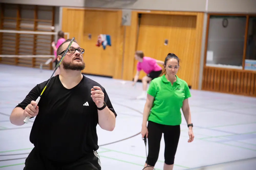 Ein Badmintonspieler in einem schwarzen T-Shirt mit Brille schaut nach oben, während er einen Federball mit seinem Schläger spielt. Im Hintergrund ist eine andere Spielerin in einem grünen Oberteil zu sehen, die ebenfalls Badminton spielt. Die Sporthalle hat einen hellen Boden und an den Wänden hängen einige Handtücher. Diese Szene könnte während des Hexennachtturniers 2026 des VfR Simmern in einem Mixed-Format aufgenommen worden sein.