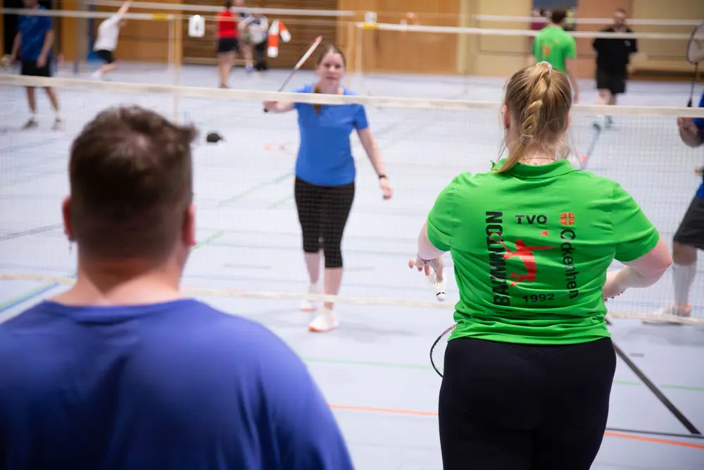 Ein Badminton-Mixed-Spiel während des Hexennachtturniers 2026 in der Halle des VfR Simmern. Im Vordergrund ist eine Spielerin in einem grünen Shirt mit dem Aufdruck „Badminton“ zu sehen, die einen Schläger hält. Im Hintergrund spielen mehrere Personen Badminton, einige in blauen und roten Shirts. Die Atmosphäre ist sportlich und dynamisch, mit Badminton-Netzen, die die Spielfelder abgrenzen.