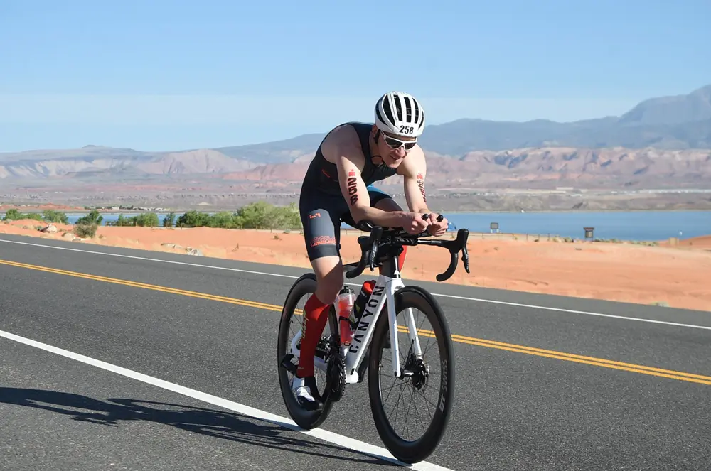 Triathlet Stefan Scherer fährt mit dem Rennrad auf einer Straße in St. George, Utah, während der IRONMAN Weltmeisterschaft 2022. Er trägt einen schwarzen und roten Wettkampfanzug mit der Nummer 258 und hat eine aerodynamische Haltung auf seinem weißen Canyon-Rennrad. Im Hintergrund sind die charakteristischen roten Felsen und der blaue Himmel zu sehen, die die Landschaft von Utah prägen.