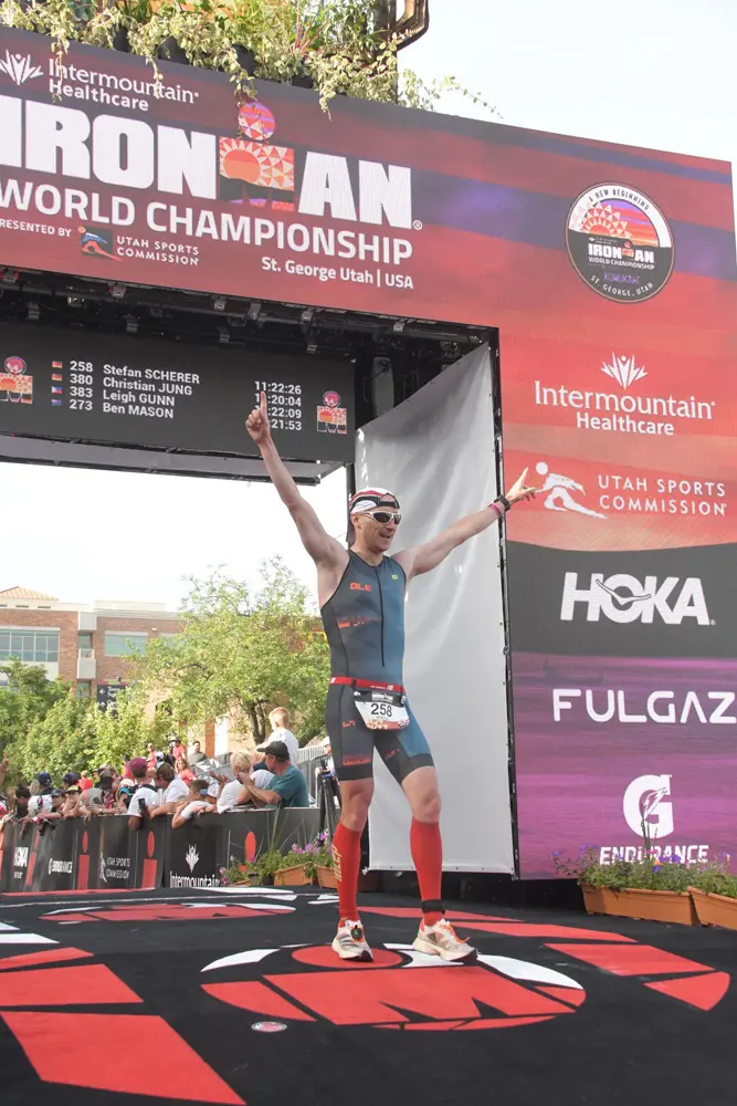 Stefan Scherer feiert seine Ankunft bei der IRONMAN-Weltmeisterschaft 2022 in St. George, Utah. Er trägt ein sportliches Triathlon-Outfit und hebt beide Arme in die Luft, während er auf dem roten Teppich des Zielbereichs steht. Im Hintergrund ist ein großes Banner mit der Aufschrift „IRONMAN WORLD CHAMPIONSHIP“ sowie eine Rangliste mit den Zeiten der Teilnehmer zu sehen.