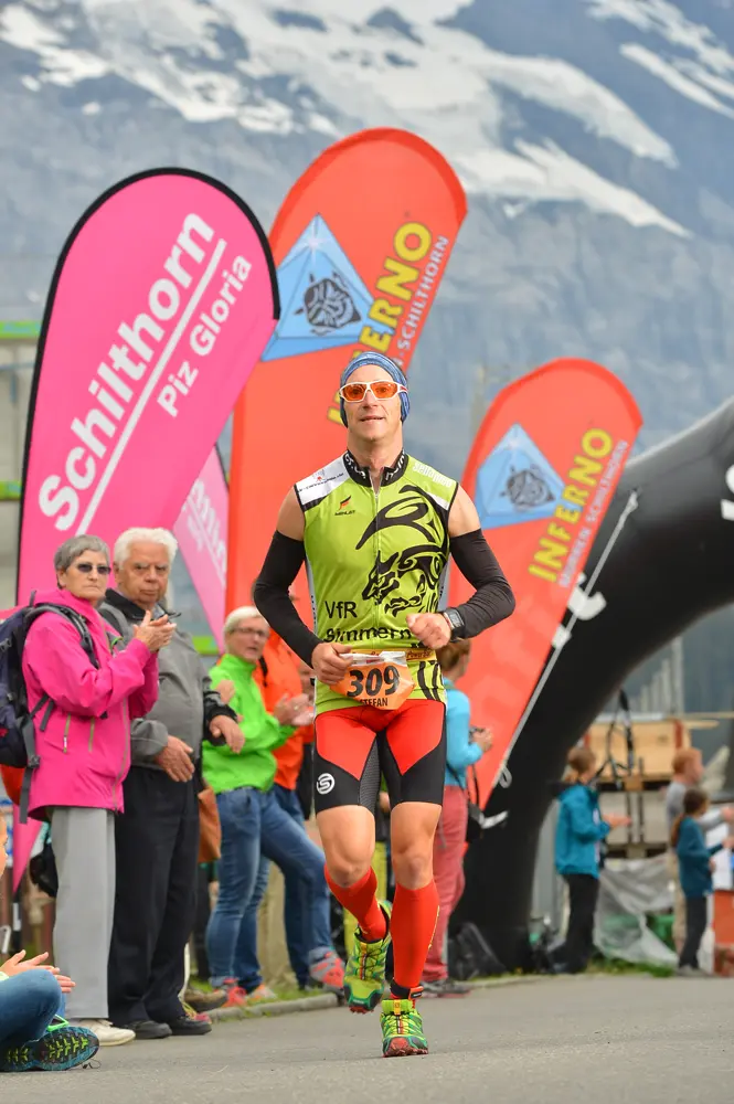 Triathlet Stefan Scherer mit der Startnummer 309, gekleidet in ein grünes und rotes Trikot des VfR Simmern, läuft in Richtung der Ziellinie beim Inferno Triathlon am Schilthorn, Schweiz. Im Hintergrund sind auffällige Werbefahnen Schilthorns und des Inferno Triathlons zu sehen, während Zuschauer ihm applaudieren.