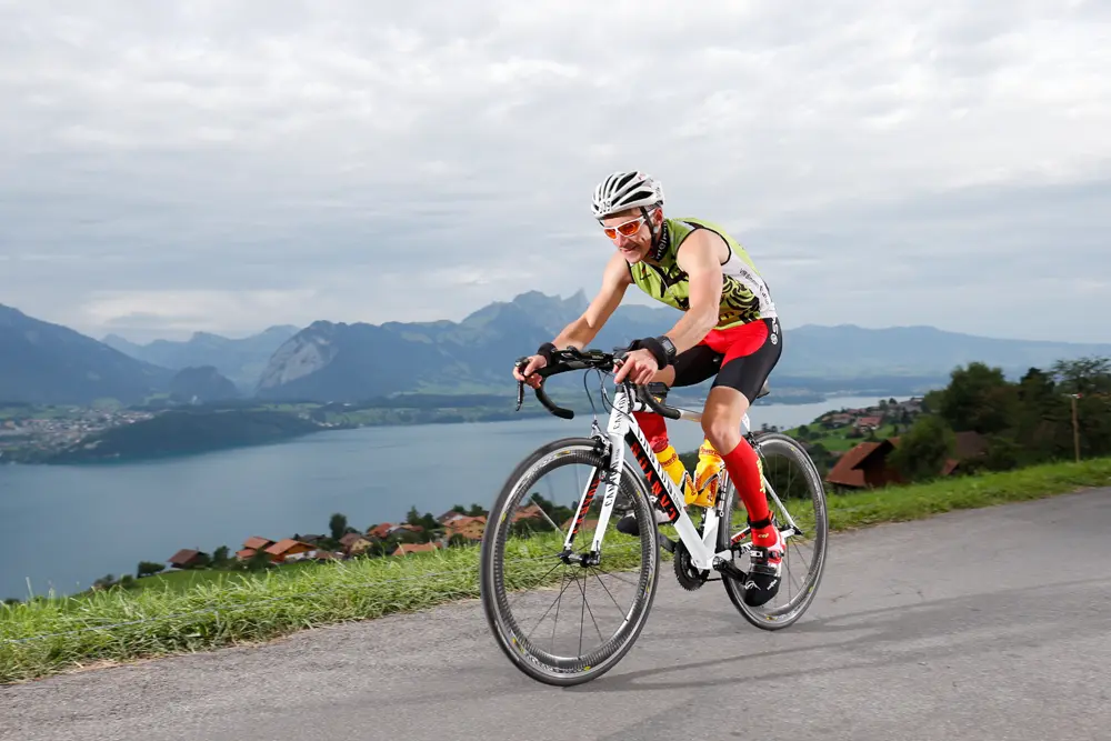 Triathlet Stefan Scherer fährt auf einem weißen Rennrad die Straße entlang, umgeben von einer malerischen Landschaft mit Bergen und einem See im Hintergrund. Er trägt ein farbenfrohes Sportoutfit mit einem grünen Oberteil und roten Shorts, sowie einen Helm und eine Sonnenbrille. Diese Szene entstand beim Inferno Triathlon am Schilthorn in der Schweiz.