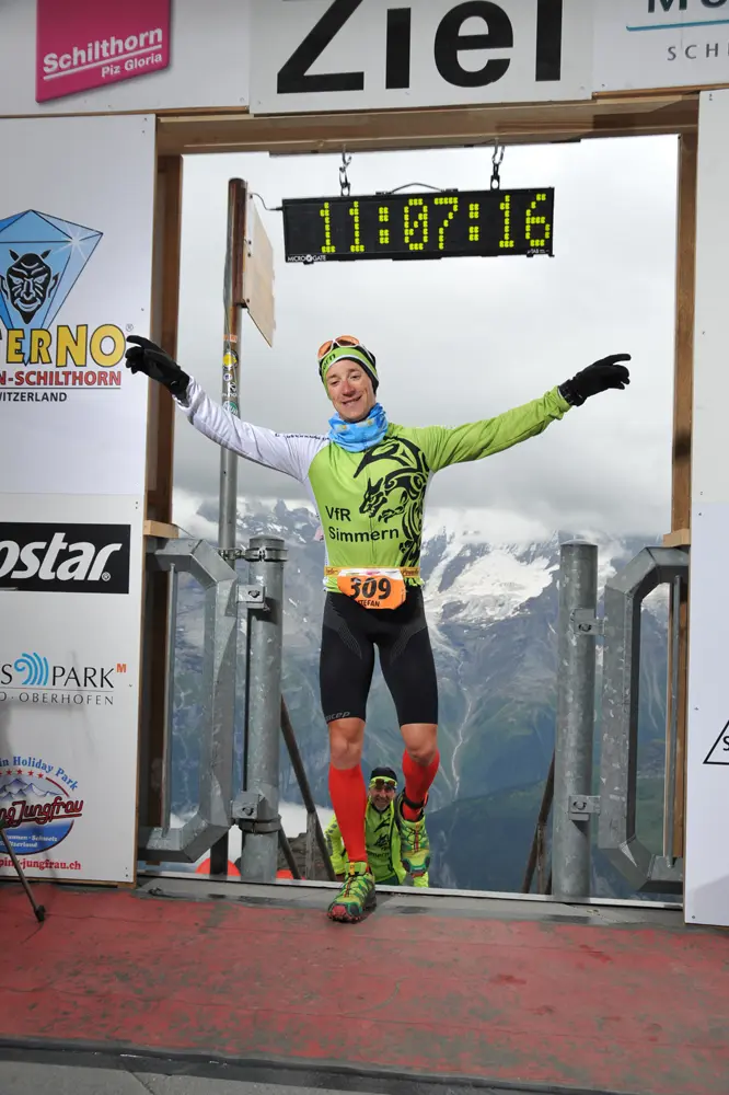 Triathlet Stefan Scherer in einem grünen Trikot mit der Aufschrift „VfR Simmern“ und der Nummer 309 feiert seinen Zieleinlauf beim Inferno Triathlon am Schilthorn in der Schweiz. Die Uhr zeigt eine Zeit von 11:07:16 an. Im Hintergrund sind Berge und eine weitere Person in grüner Sportkleidung zu sehen.